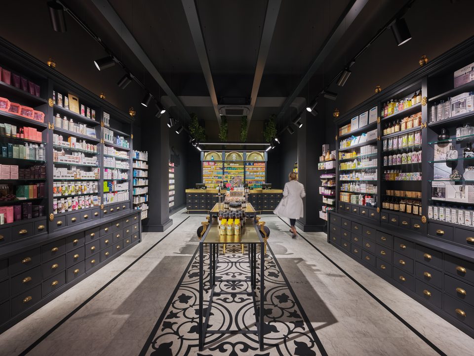 Frontaler Blick durch die dunkle Offizin der Apotheke Schwarzer Adler in Wien, zwischen den Schubladenschränken führt der Ornamentboden bis zur golden beleuchteten Rückwand