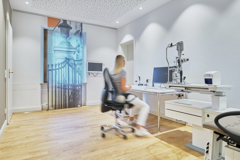 Untersuchungsraum im Altbau: eine Mitarbeiterin am Arbeitsplatz zwischen den augenärztlichen Geräten, an der Wand ein raumhoher Fotodruck des schmiedeeisernen Schlosstors