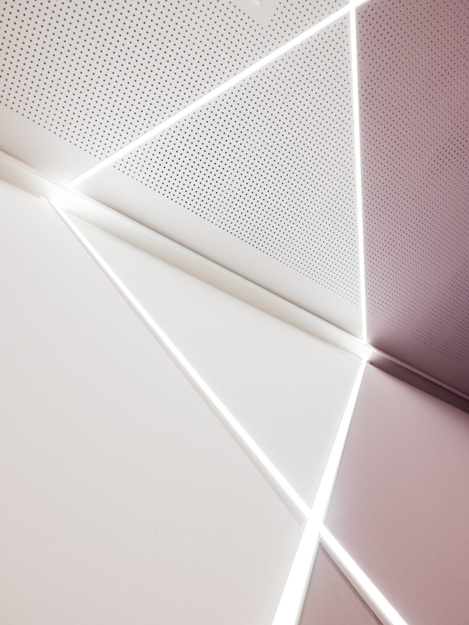 Grafisches Detail der Deckenecke, in der sich drei LED-Linien vor perforierter Akustikdecke und roséfarbener Fläche kreuzen