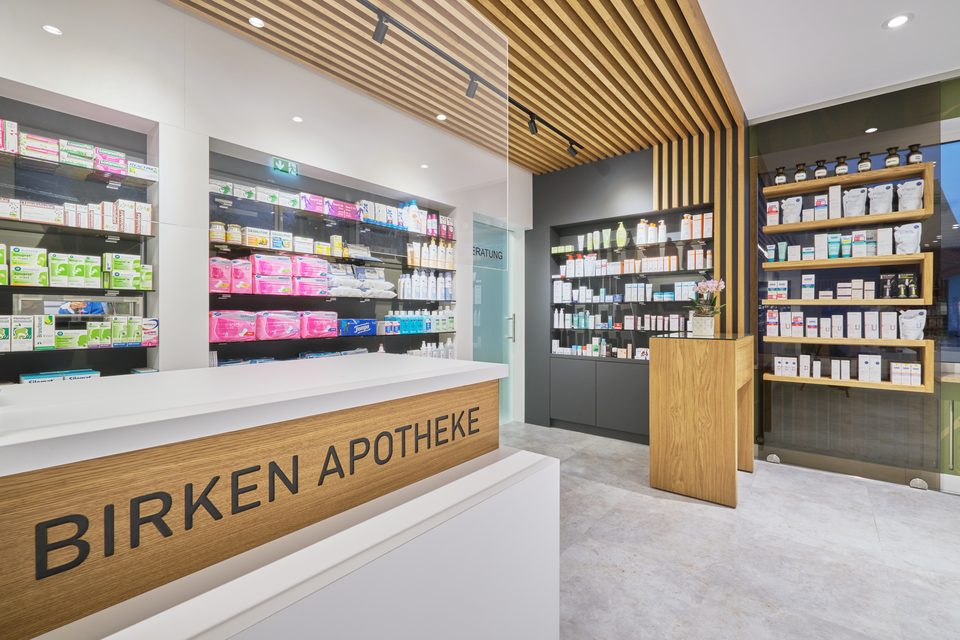 Handverkaufstresen der Birkenapotheke, auf dessen Holzband der Schriftzug „Birken Apotheke“ steht, dahinter die Sichtwahl unter der Lamellendecke