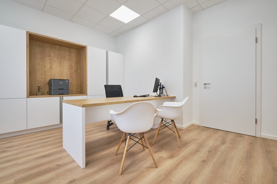 Arztzimmer mit Schreibtisch aus hellem Holz, weißem Stuhl und Einbauschrank