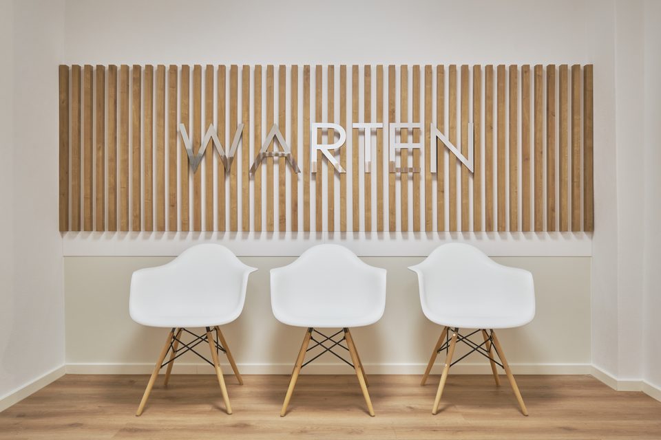 Wartebereich mit dem Wort „Warten“ in Metalllettern auf der Holzlamellenwand, darunter drei Schalenstühle