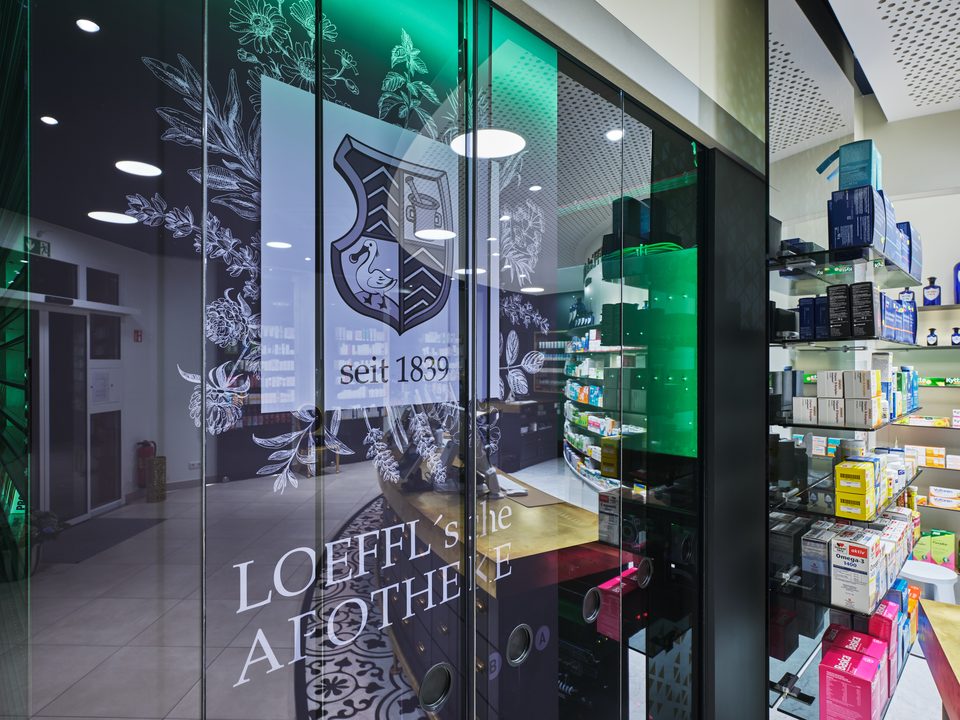 Weite Offizin der Löffl’schen Apotheke Schörfling mit dunkler Decke und beleuchteter Sichtwahl