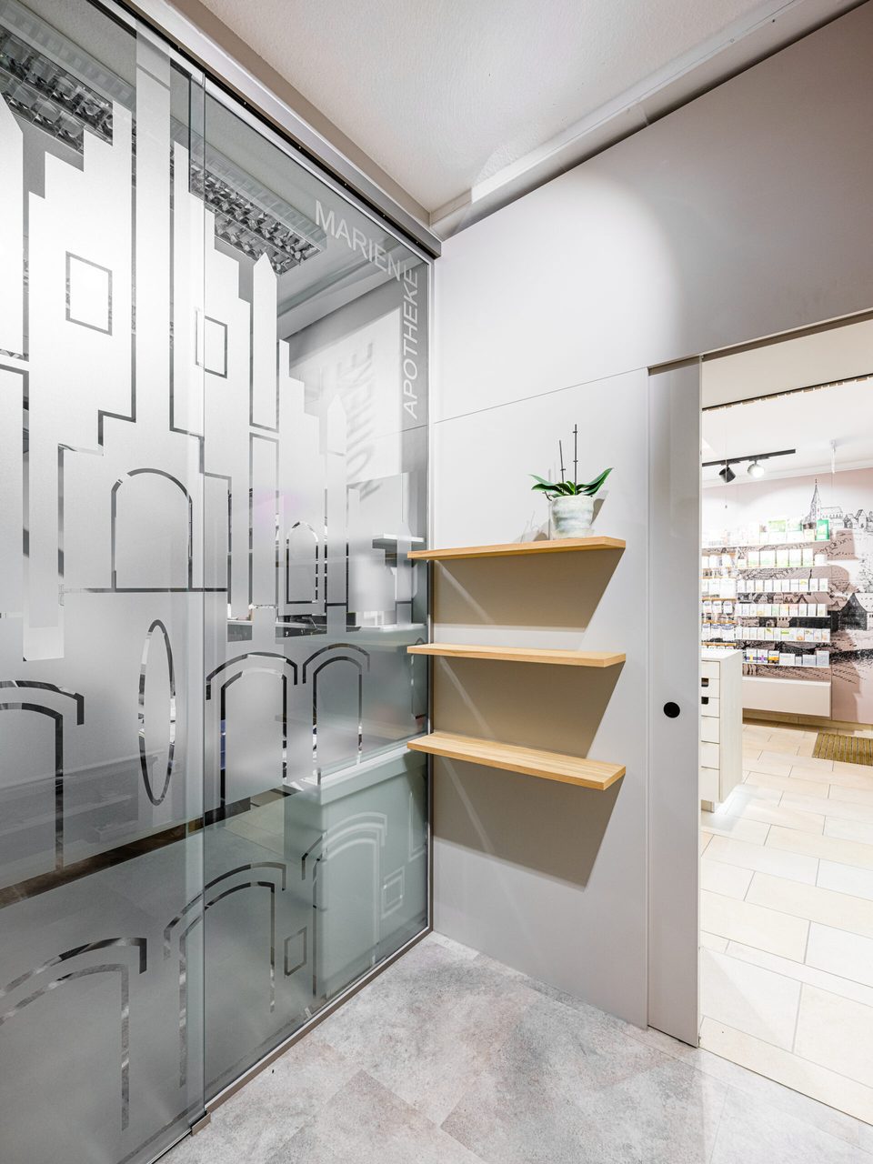 Satinierte Glaswand mit eingeätzten Umrissen der Freisinger Bauten, rechts daneben schwebende Holzregalböden mit einer Orchidee