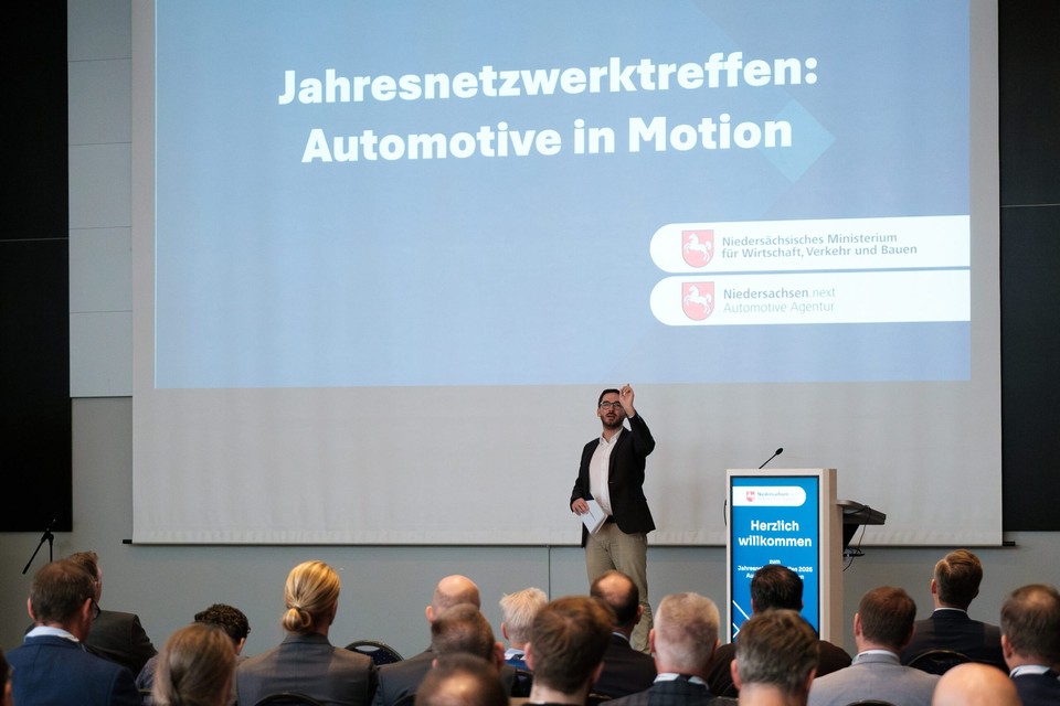 Begrüßung am Pult vor der Leinwand mit dem Leitthema „Jahresnetzwerktreffen: Automotive in Motion“.
