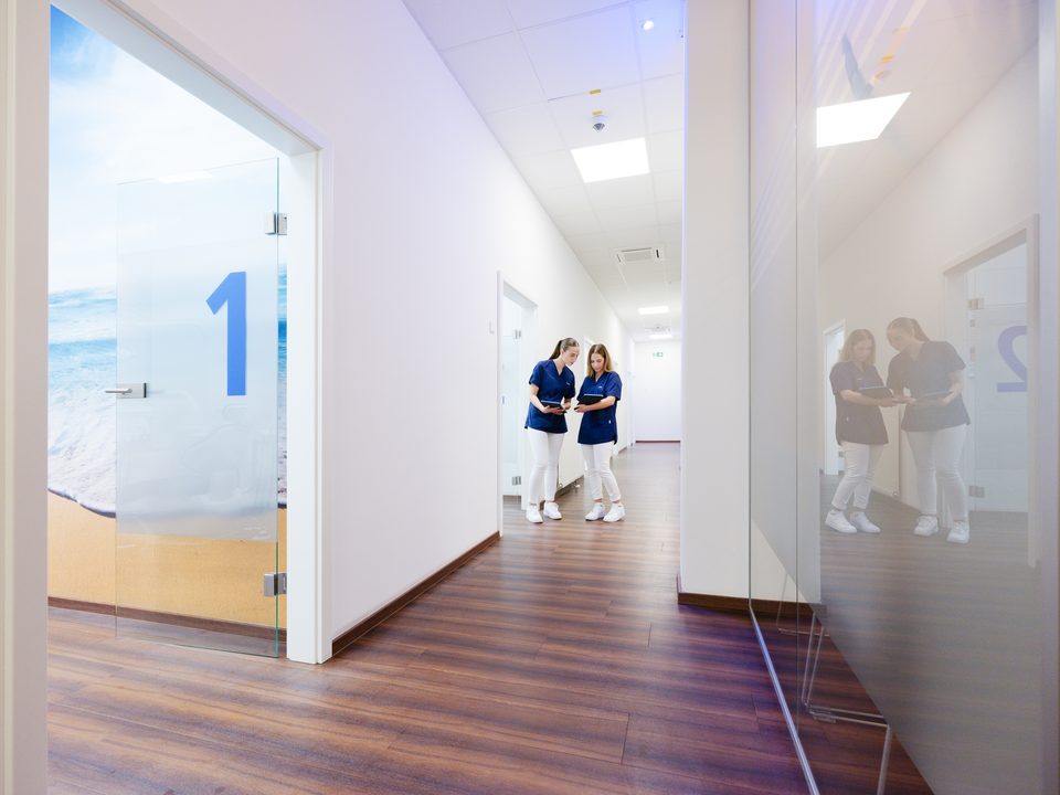 Flur mit Glastrennwänden, deren blaue Ziffern die Behandlungszimmer nummerieren, hinten das Team